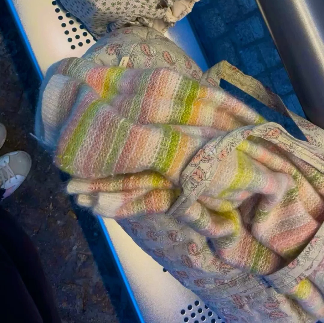 Ein bunt gestrickter Mohair Pullover in Grün- und Rosatönen, liegend auf einer Reisetasche auf einer Bank
