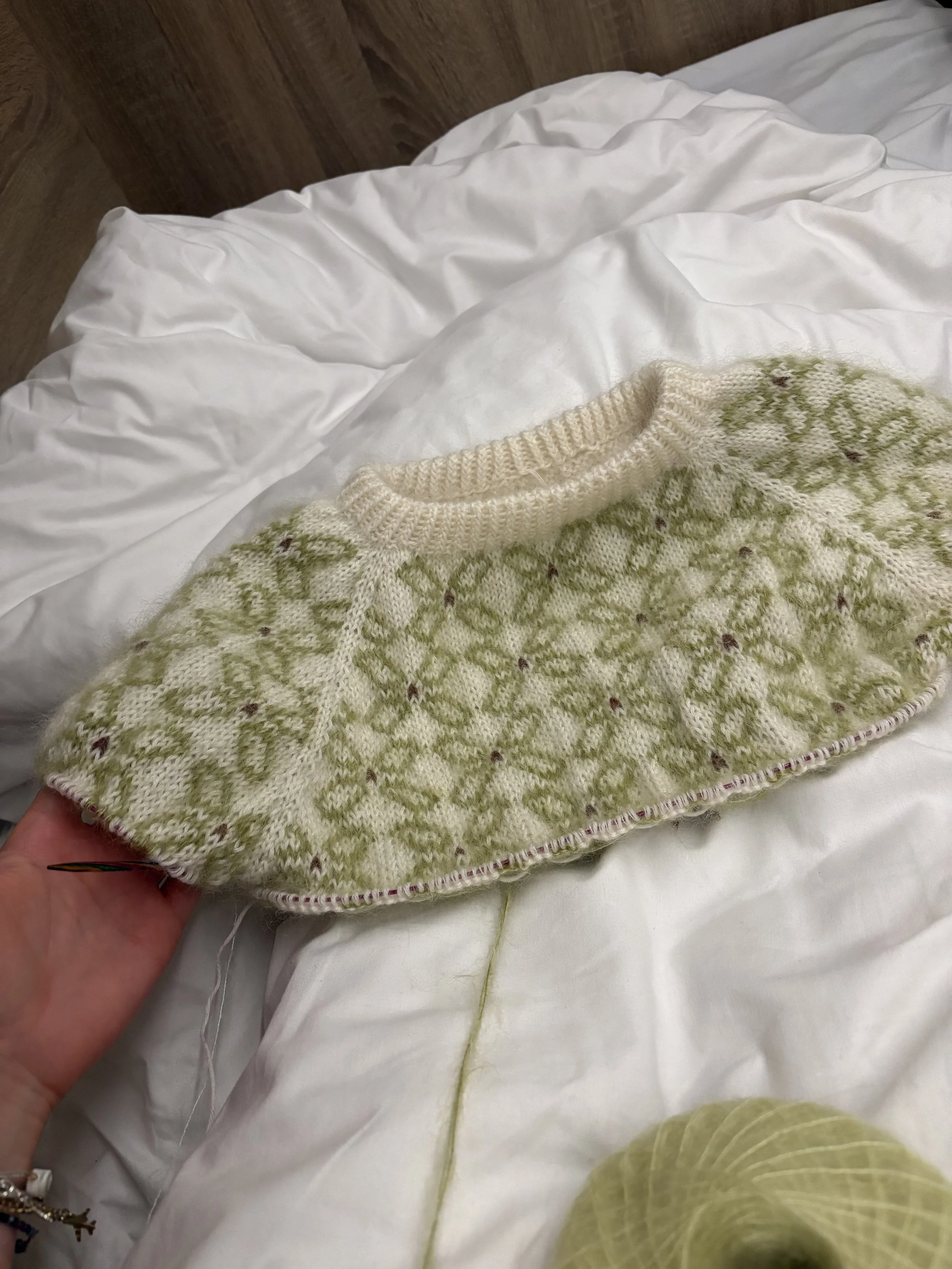 Ein Weiß Grüner Fair-Isle-Pullover mit Hand gestrickt mit Blumenmuster fotografiert in einem Bett mit Weißen Laken.