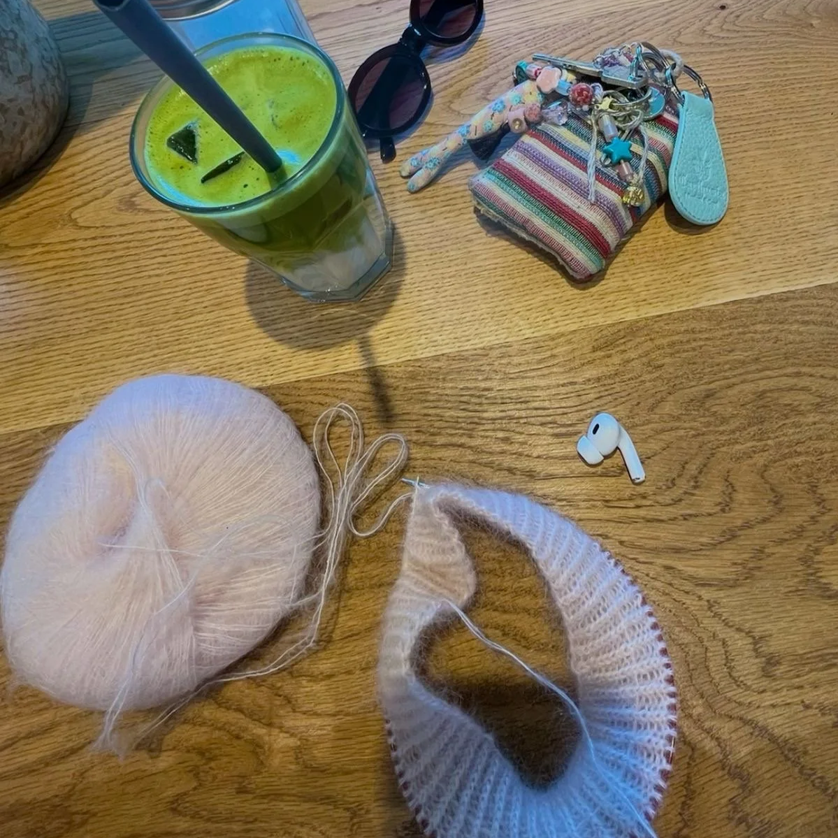 Holztisch in einem Café mit einem Iced Matcha, einem Strickprojekt aus rosanem Mohair, Sonnenbrille und Kopfhörer