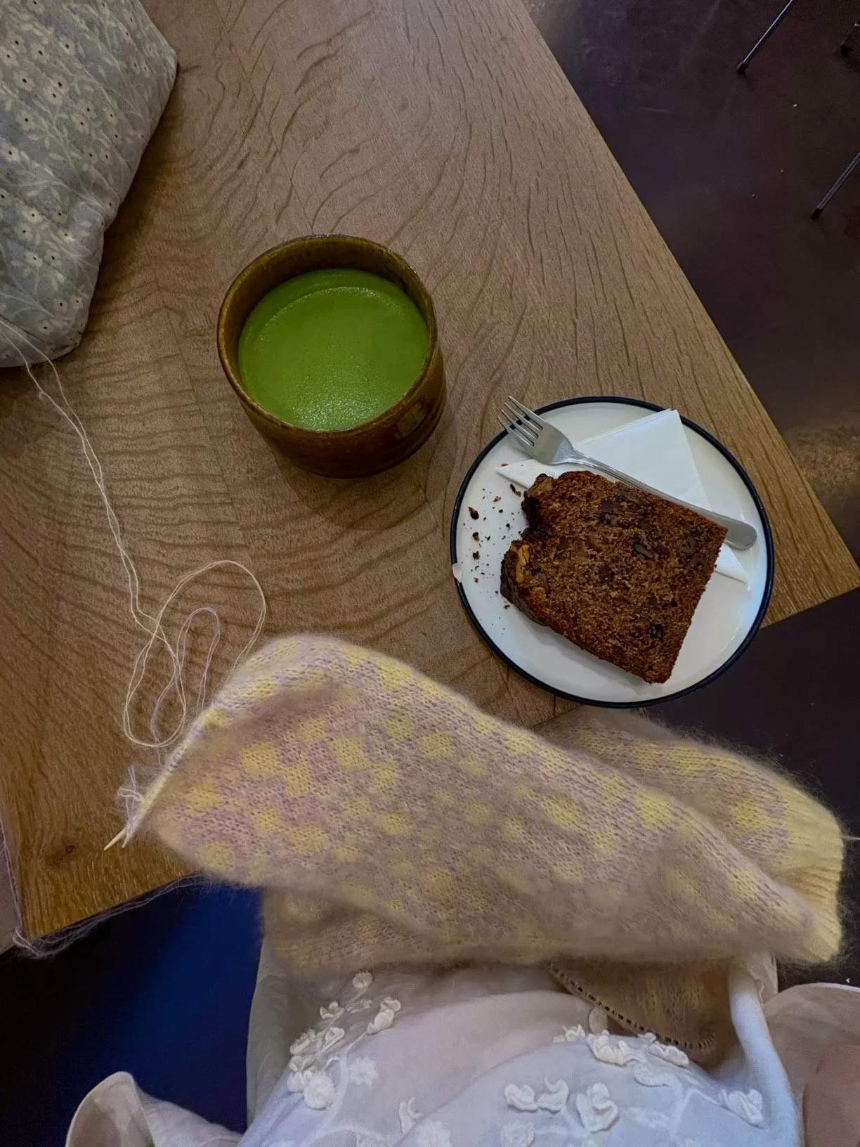 Holztisch in einem Cafe mit einer Tasse Matcha, einem Teller mit Bananenrot und ein Strickprojekt in gelb lila Fair-Isle-Muster 