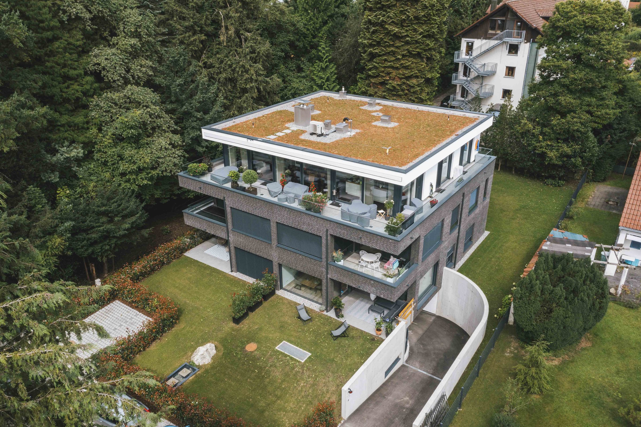 Modernes Mehrfamilienhaus mit Balkon im Garten, umgeben von Bäumen, mit Dachbegrünung und Terrassenmöbeln.