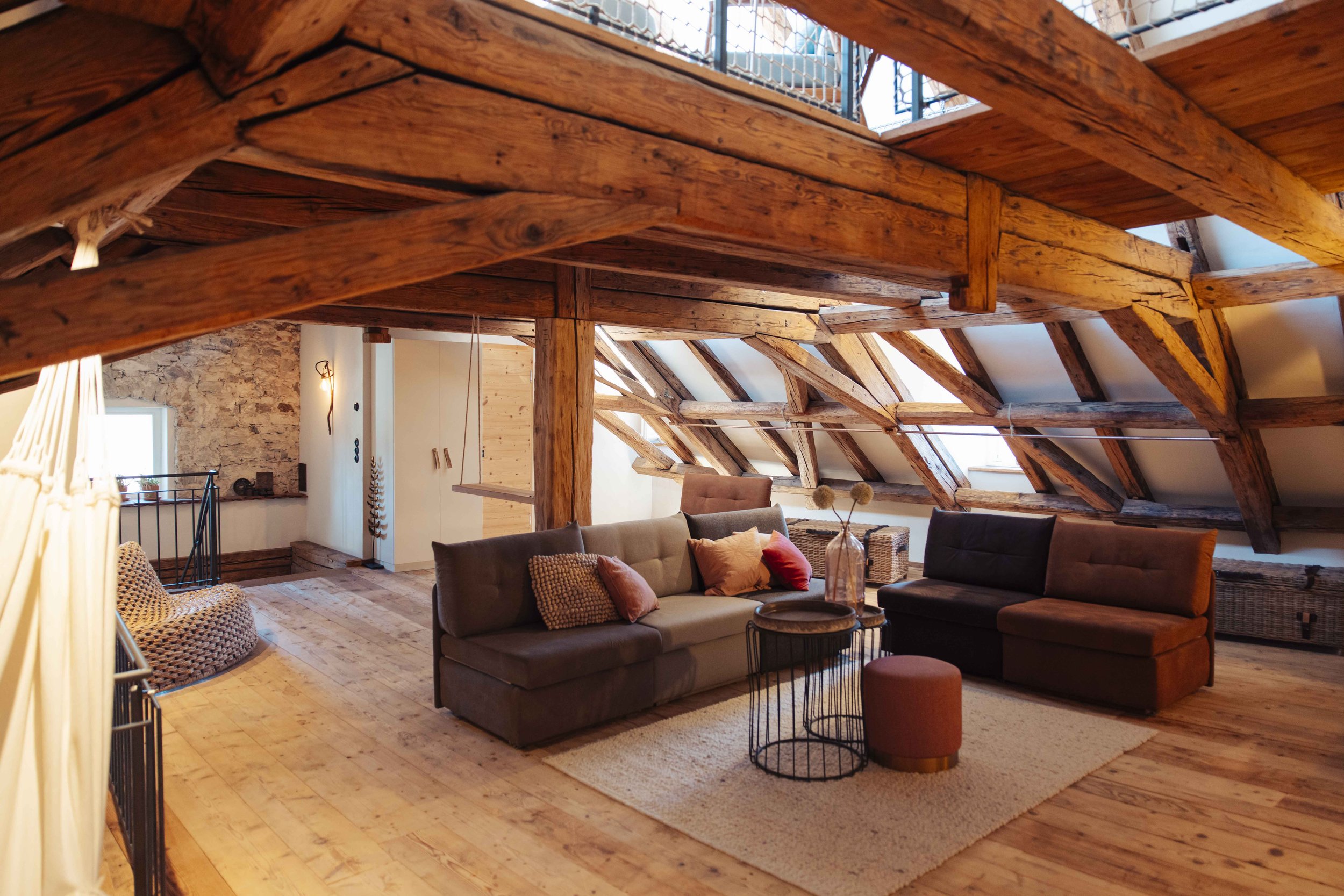 Gemütliches Wohnzimmer mit großen Holzbalken und Holzdecke, bequemer Couchecke, Teppich, seitlicher Fenster, verspiegelter Boden, indirekte Beleuchtung.