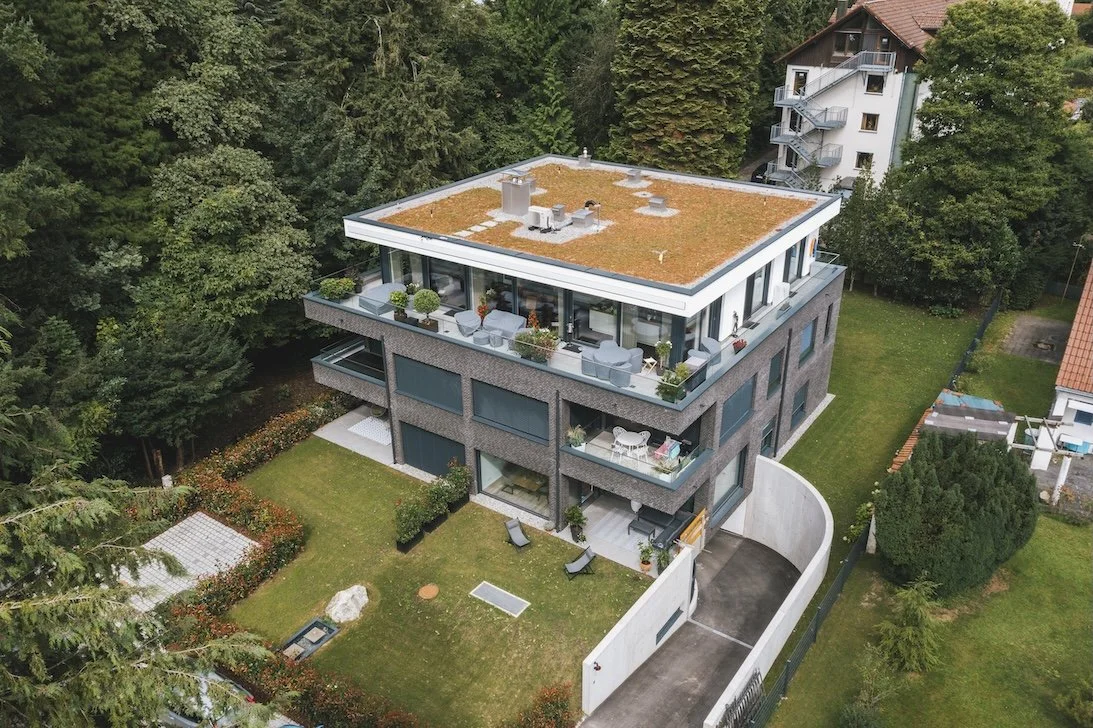 Mehrstöckiges Haus mit großen Terrassen, umgeben von Bäumen und grünem Garten, Dach mit Begrünung, auf einer Straße in einer Wohngegend.