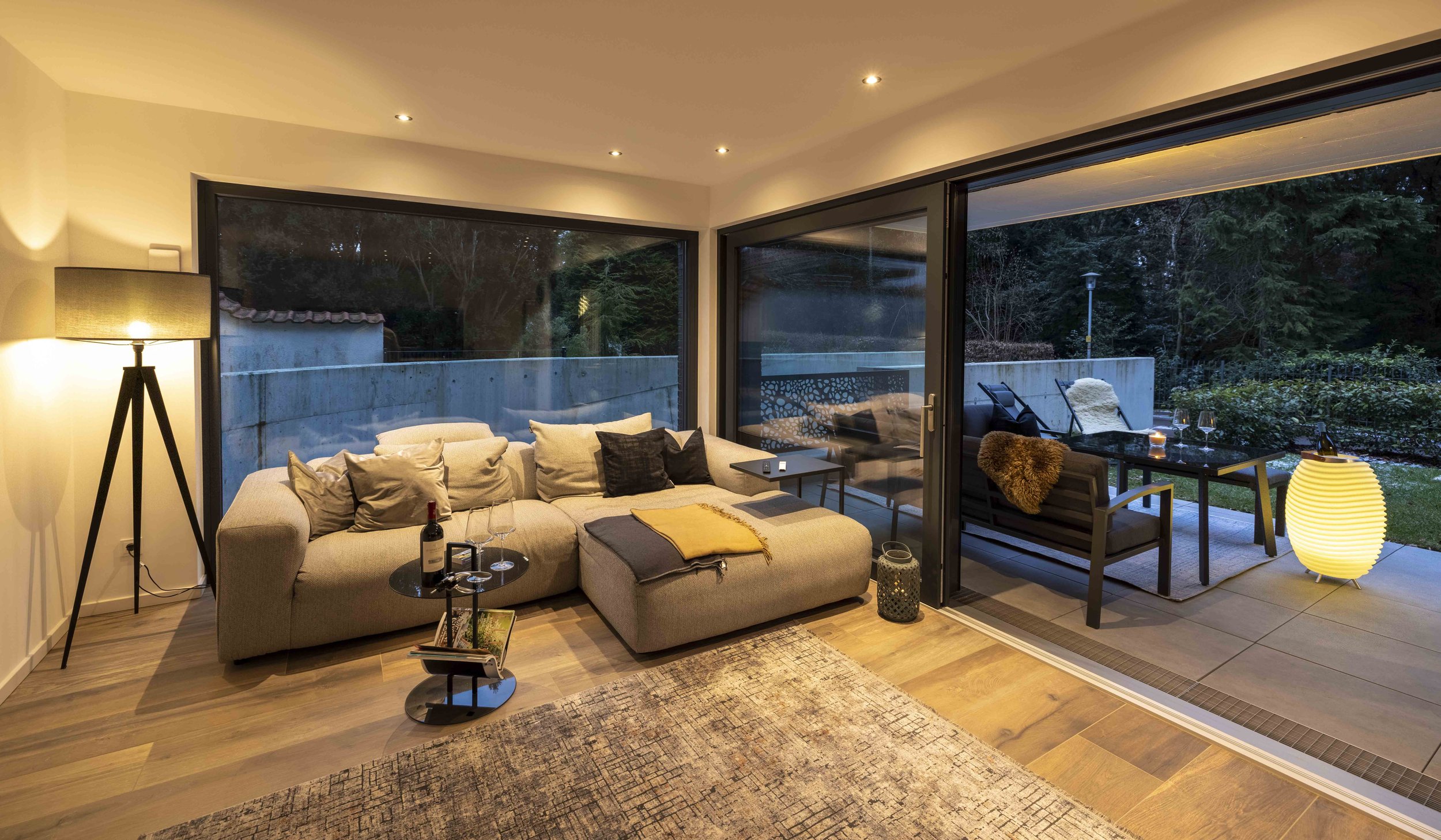 Gemütliches Wohnzimmer mit großen Fenstern und Aussicht auf den Garten, eingerichtet mit einem großen Sofa, einem Beistelltisch, Stehleuchte und einer Terrasse mit Gartenmöbeln, bei Nacht