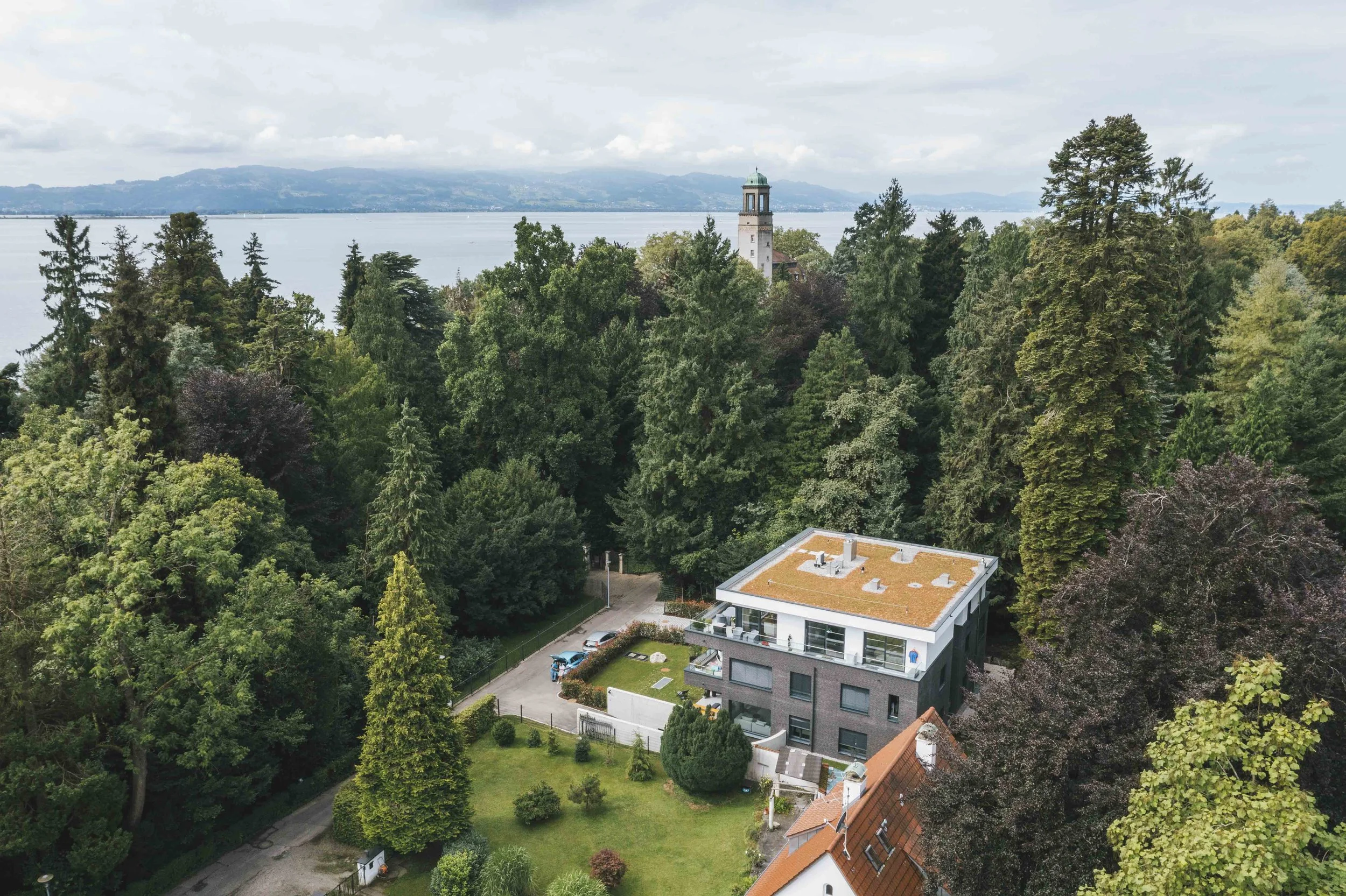 Ein Haus mit weißen und grauen Wänden, rotem Dach und großen Fenstern, umgeben von einem grünen Garten und umrahmt von hohen Bäumen. Im Hintergrund sieht man einen See mit Bergen. 