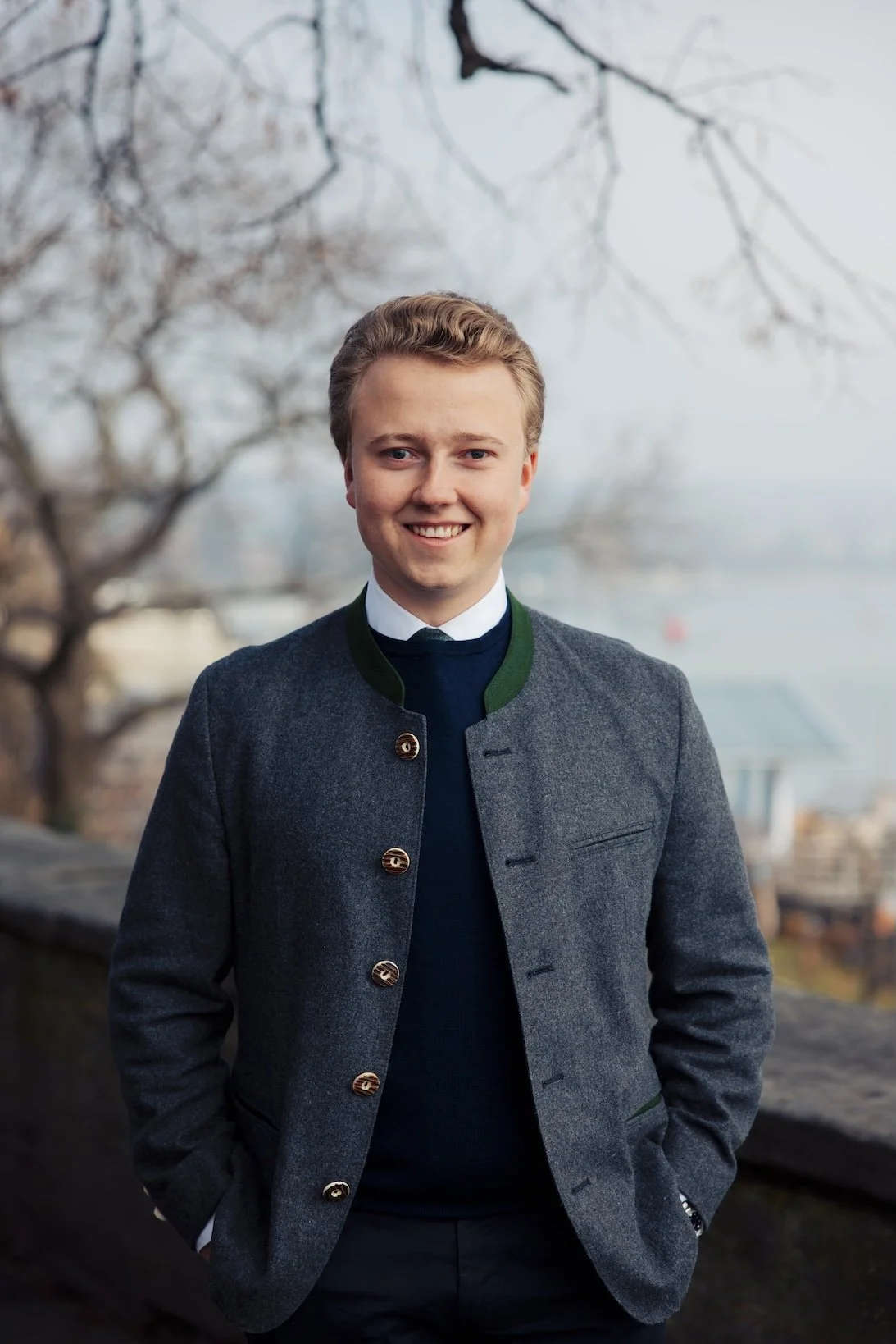 Ein lachender junger Mann in einem grauen Anzug mit grünem Kragen steht vor einer Baumlandschaft mit Wasser im Hintergrund.