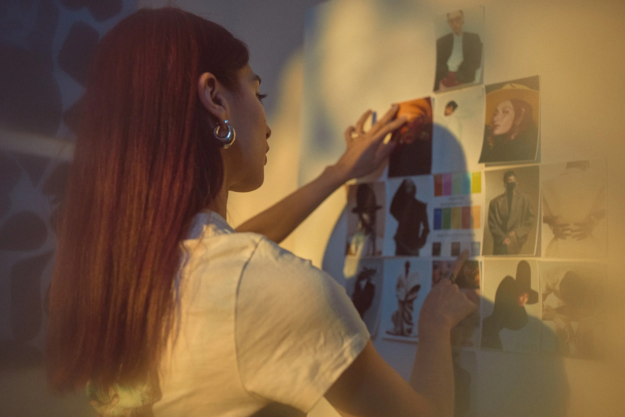 A woman with long red hair and hoop earrings is reaching towards a wall decorated with various photographs and color pattern charts. She is wearing a light-colored top.