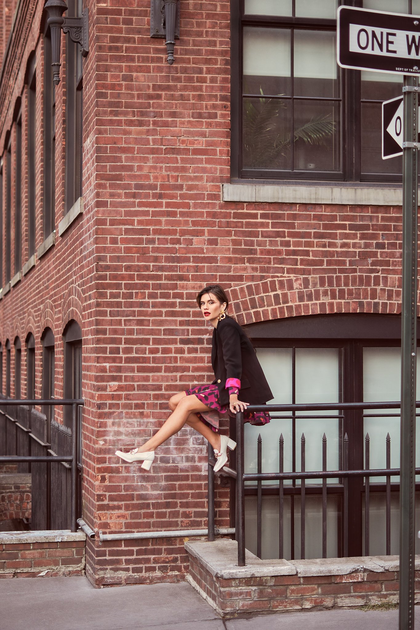 A woman with dark hair, wearing a black blazer, pink leopard print dress, and white high heels, sitting on a black metal railing outside a red brick building with a large window and a 'One Way' street sign.