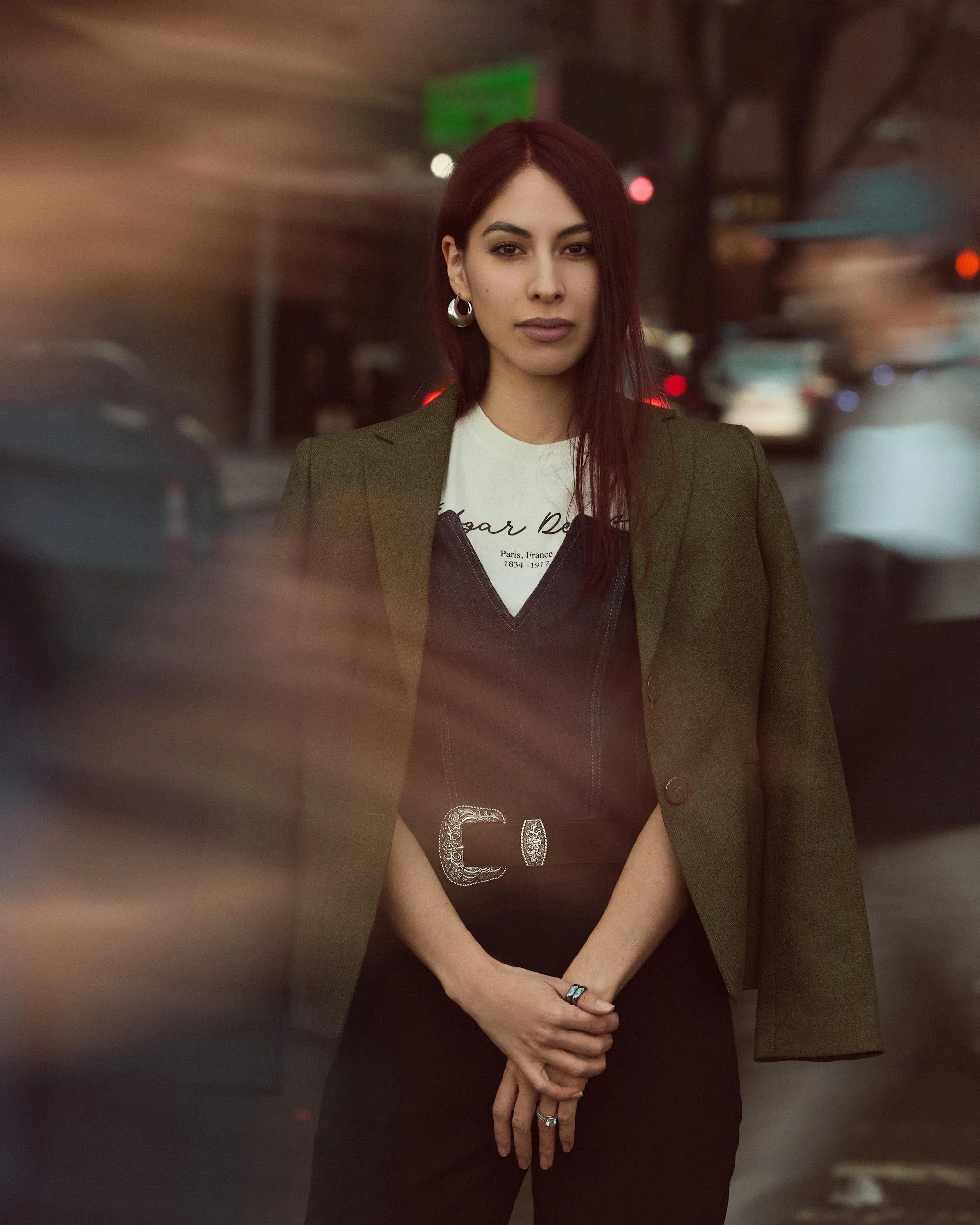 A woman with long red hair and hoop earrings, wearing a white t-shirt and an olive-green blazer, standing in a city street with blurred cars and streetlights in the background.