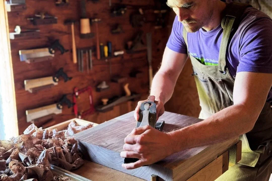 A man in a purple T-shirt and work apron is smoothing a piece of wood with a hand plane in a woodworking shop, with woodworking tools hanging on the wall in the background.