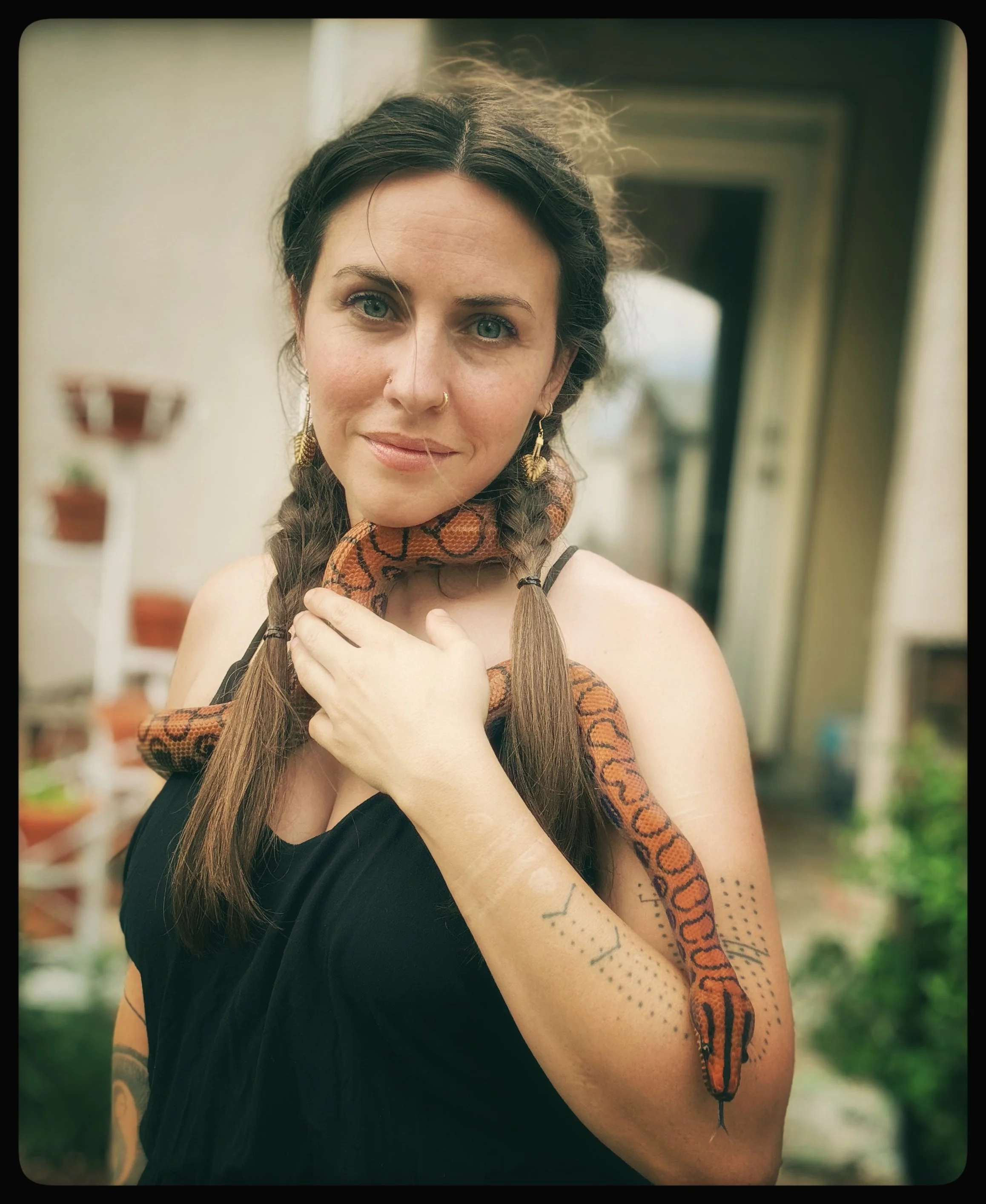 A young woman with long brown hair and blue eyes, wearing a black top, earrings, and a patterned scarf, holding a snake around her shoulders and arm with tattoos on her arm, standing in front of a blurred outdoor background.
