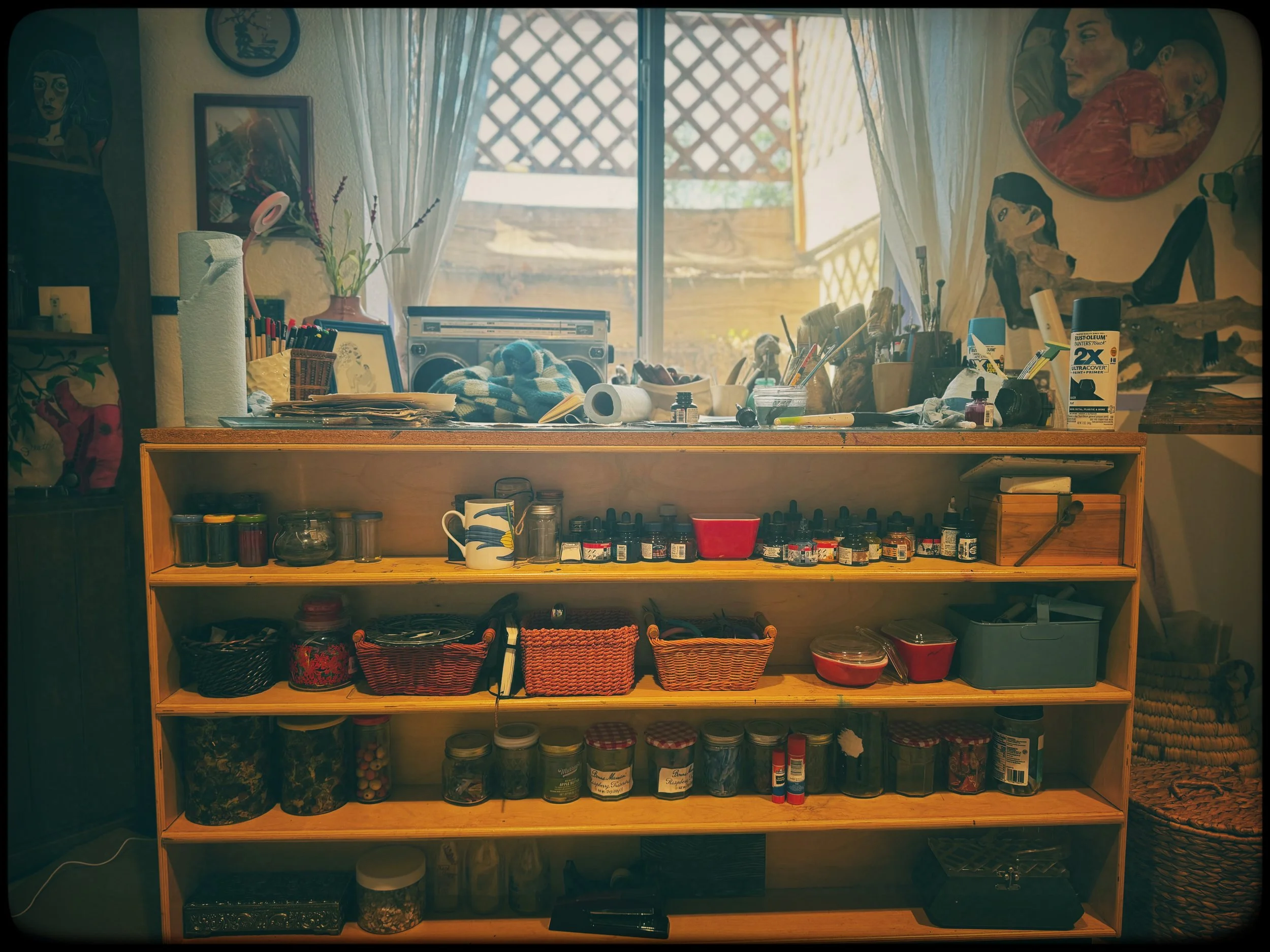 A cluttered wooden shelf filled with art supplies, jars, containers, and baskets, with a window behind and paintings on the wall.