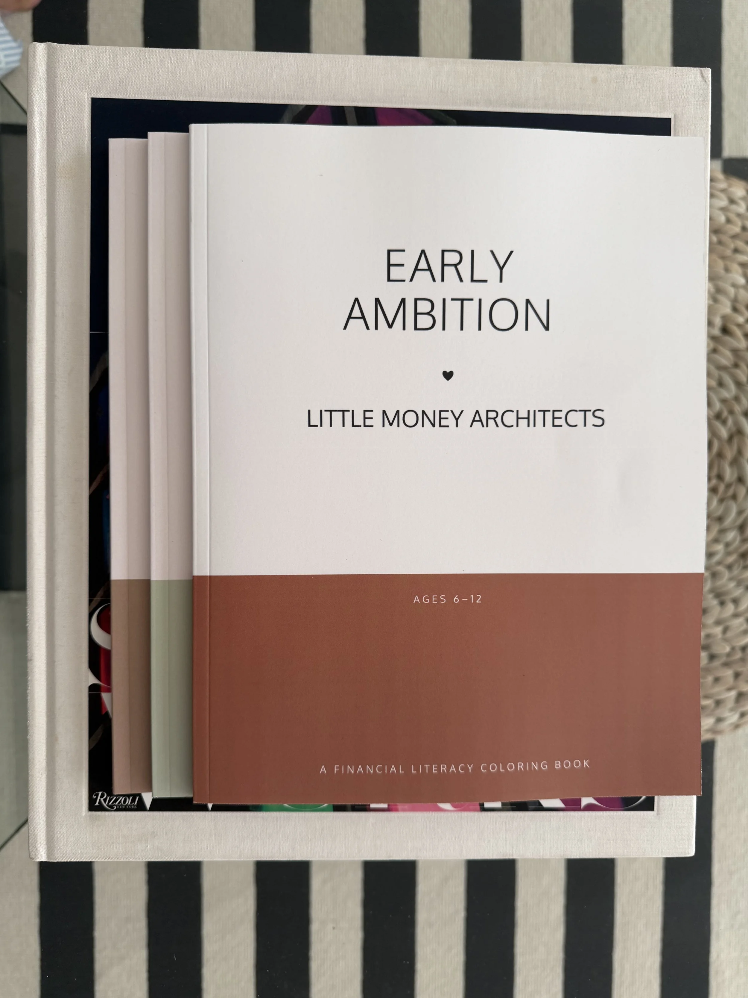 A financial literacy coloring book titled 'Early Ambition Little Money Architects, designed for ages 6-12,  placed on a white surface with other books partially visible behind it. The cover is white with a brown bottom section and black text.