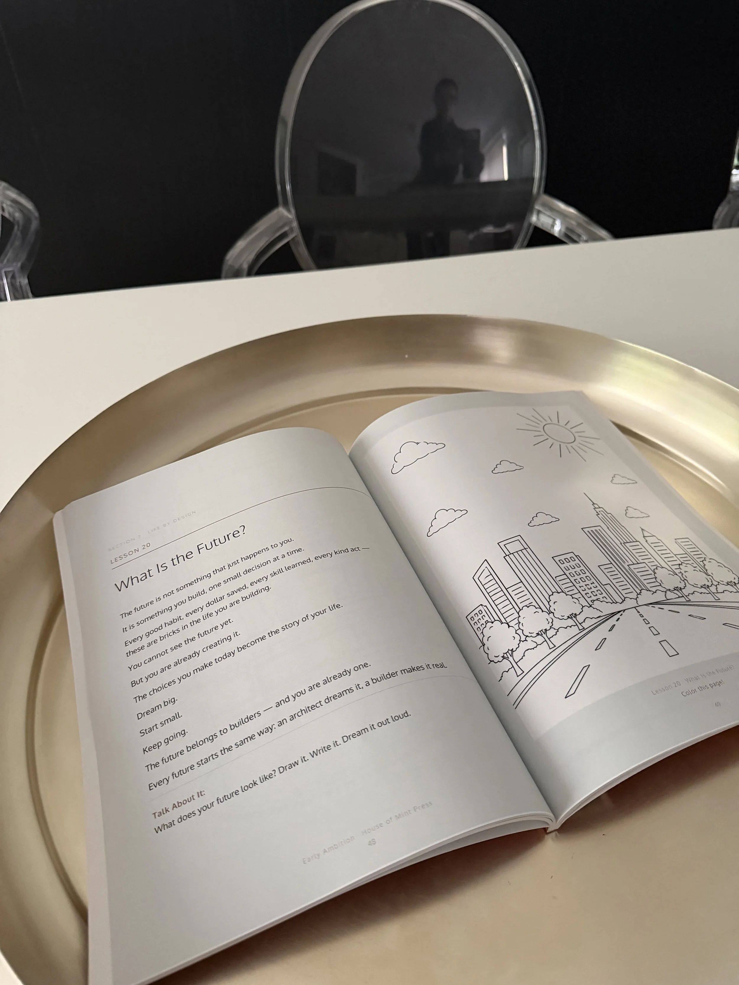 Open Early Ambition Little Money Architects financial literacy coloring  book  showing a lesson with text about the future and a black-and-white cityscape illustration with tall buildings, trees, and a sunny sky.