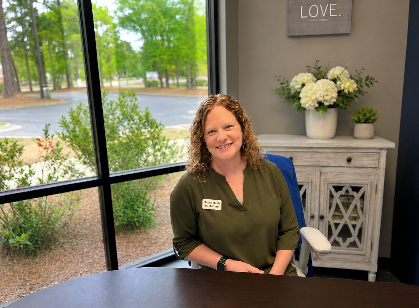 A woman with curly red hair sitting at a desk near a window, smiling, wearing a green blouse with a name tag that says 'Maureen Minds Publishing', in an office with a vase of white flowers and a framed art piece that reads 'LOVE' on the wall behind her.