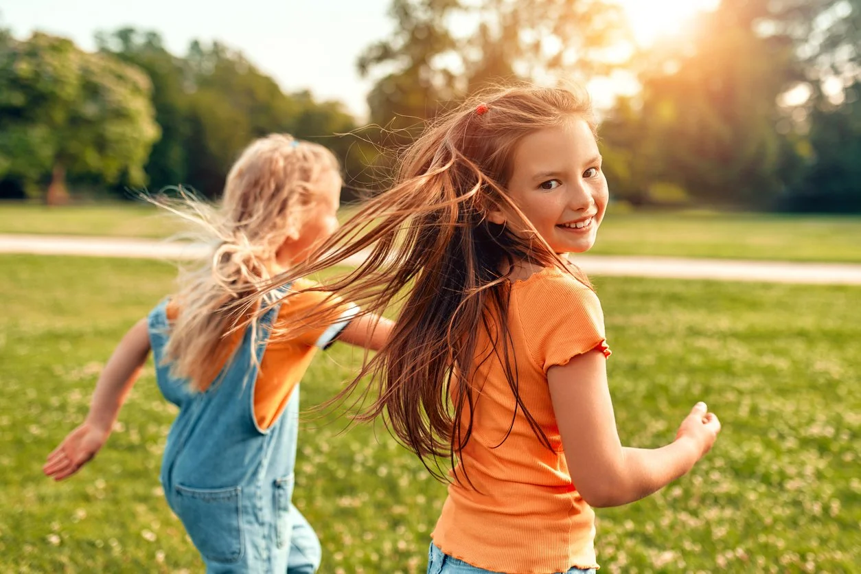 Two young girls running and smiling in a sunny park, wearing casual clothes.