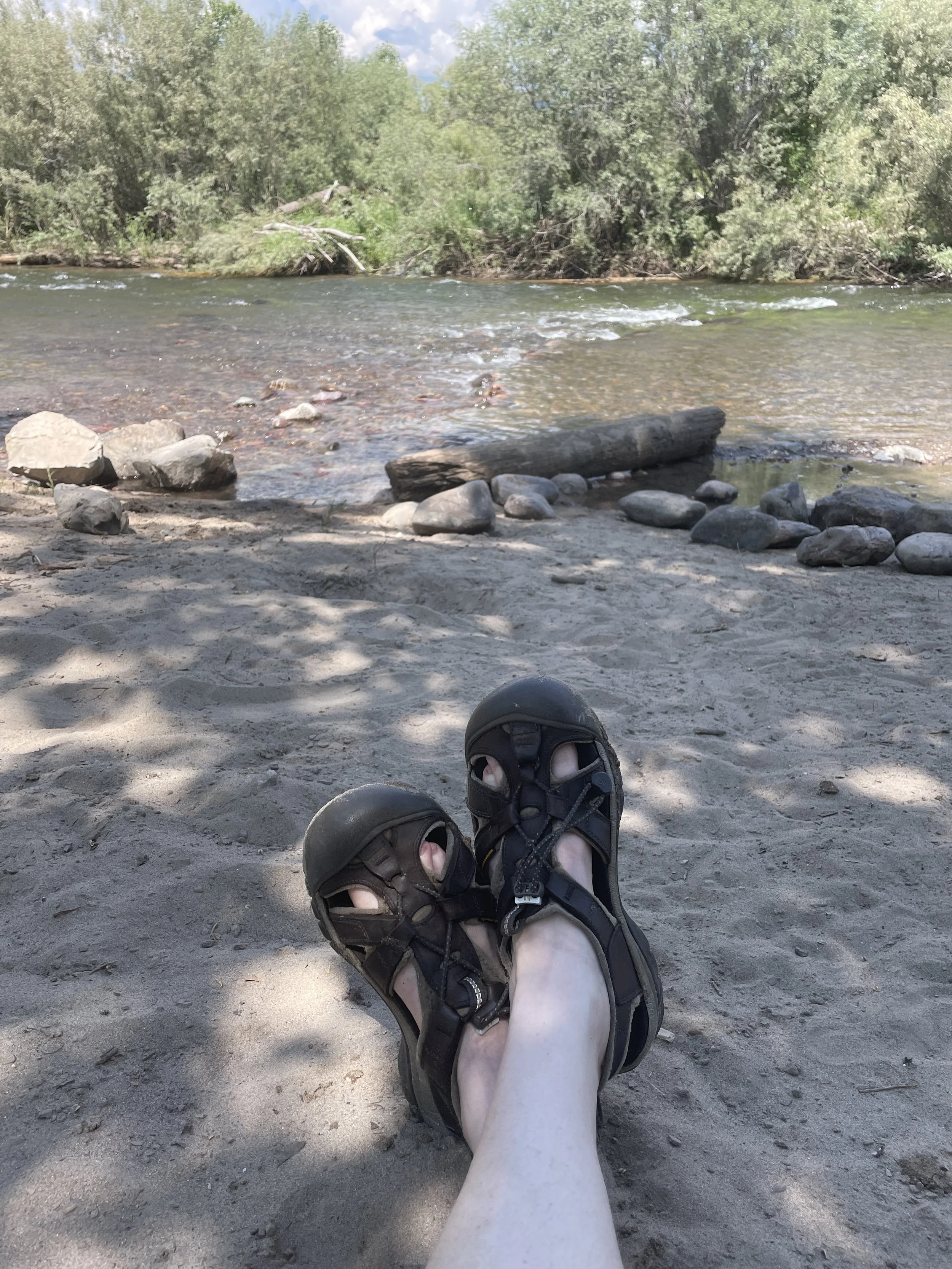 Person relaxing on a sandy riverside with their feet in sandals, overlooking a flowing river with rocks and lush green trees in the background.