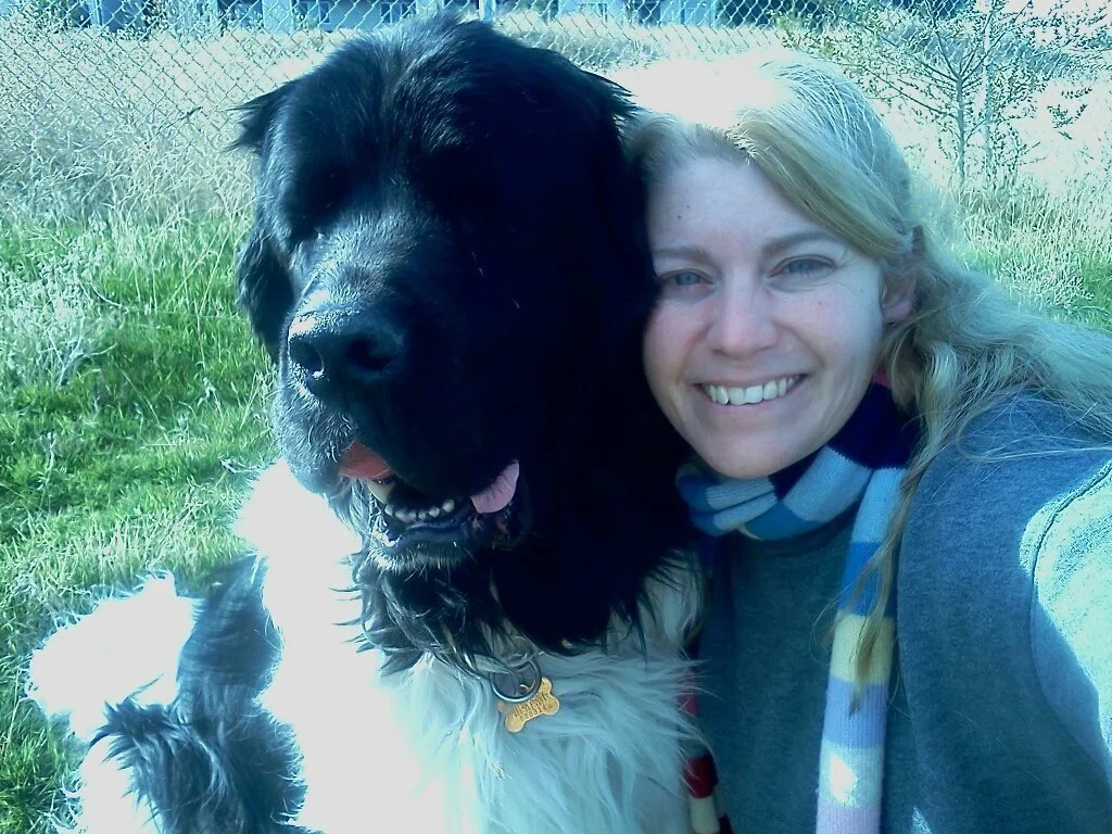 A woman smiling next to a large black and white dog outdoors on grass.