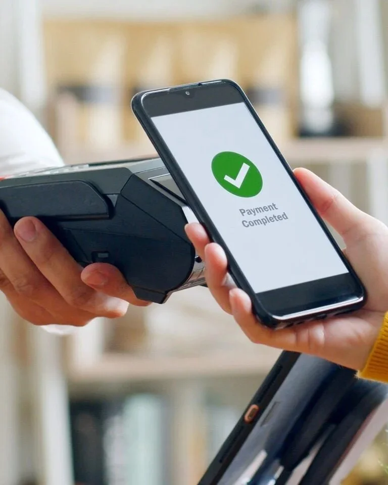 Person using a smartphone to show a payment confirmation with a large green checkmark and the words "Payment Completed" on the screen, while holding a card reader.