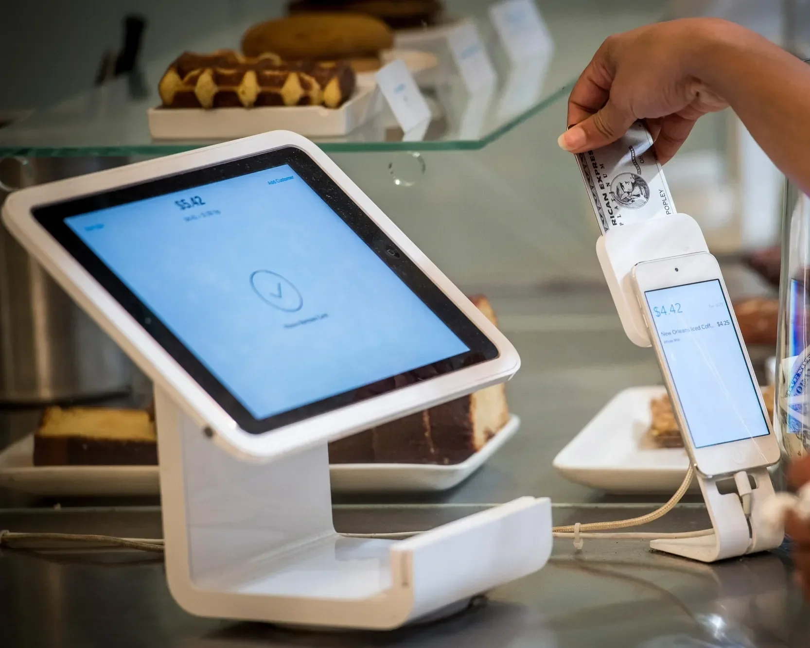 A person using a mobile payment terminal to scan a debit card at a bakery or cafe. In the background, there are trays of baked goods, including cookies and cakes, displayed on shelves.