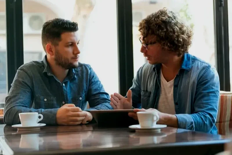 Two men are sitting at a table in a cafe having a conversation. One man is holding a tablet, and they are discussing something. Two cups of coffee are on the table, and large windows with a view of the street are behind them.