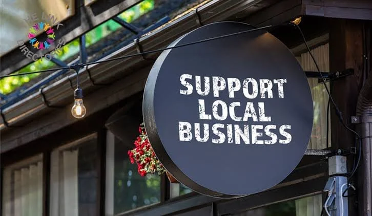 A round black sign hanging outside a building with the message "Support Local Business" written in white, with a string of light bulbs hanging above.