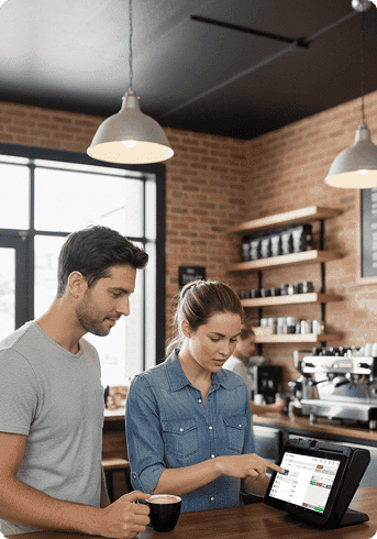 A man and woman working at a coffee shop counter with a digital register, coffee brewing equipment, and shelves with cups and supplies in the background.