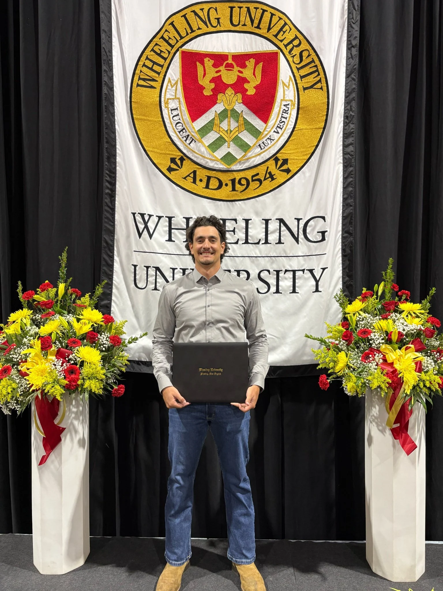 A young man standing in front of a University of Wheeling banner, holding a diploma, with colorful flower arrangements on either side of him.