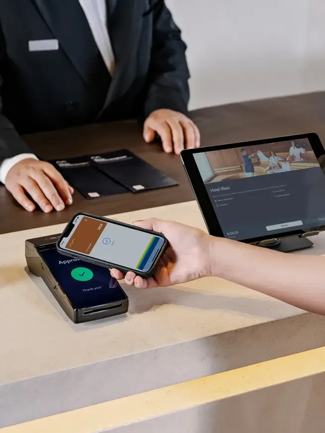 Person using a smartphone to pay at a hotel front desk with a contactless payment terminal, while a hotel employee waits behind.