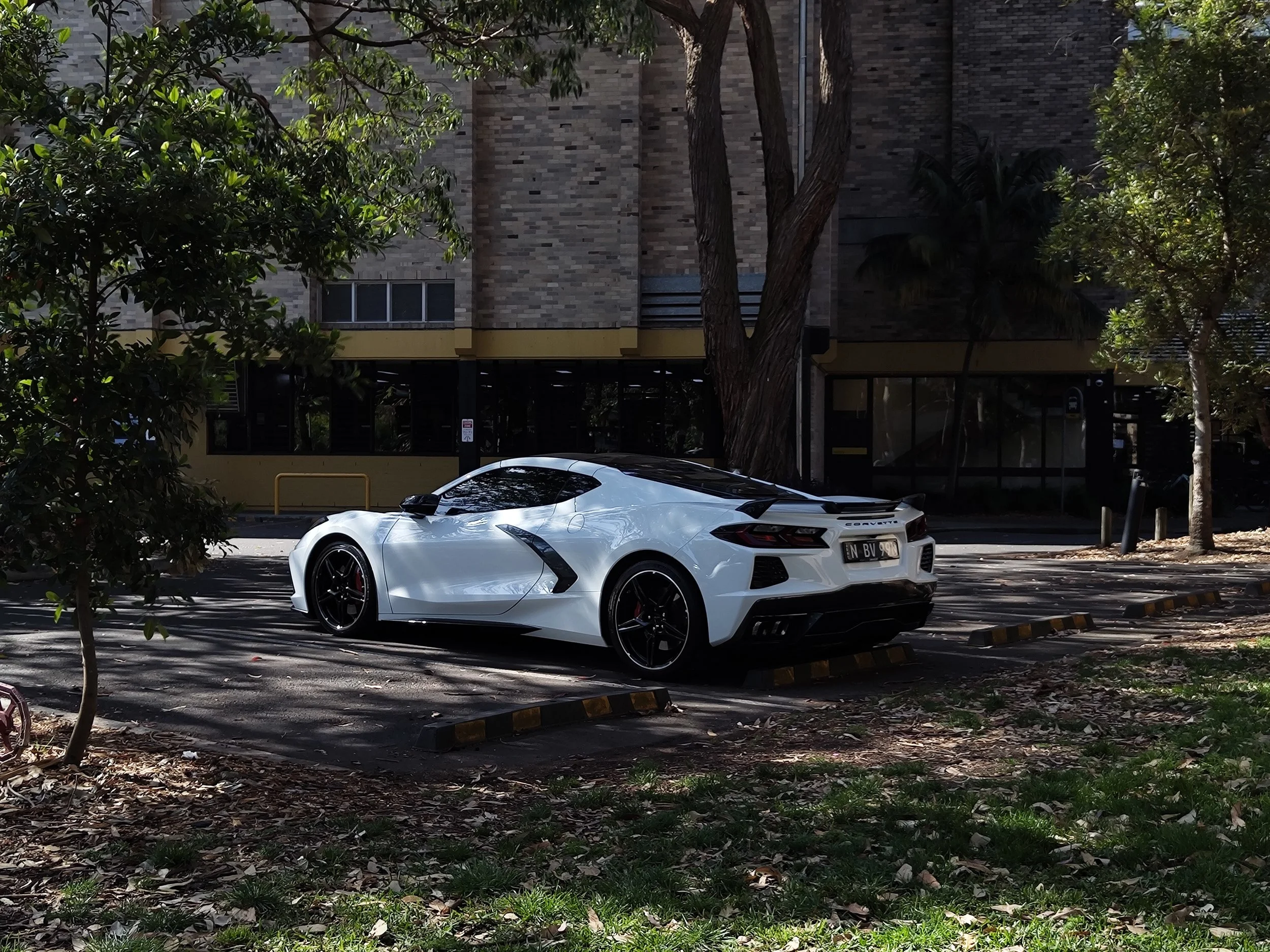 A white sports car parked in a parking lot surrounded by trees and greenery, with a building in the background.
