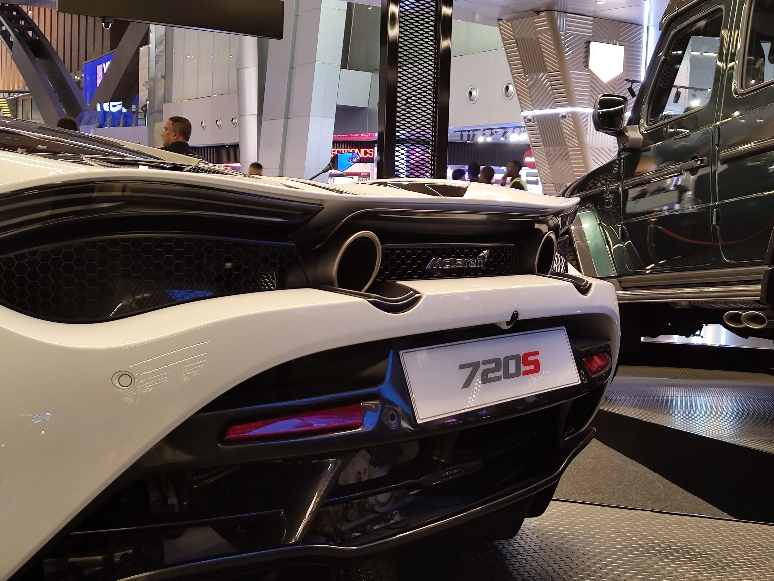 A white McLaren 720S sports car with black accents on display at an indoor auto show, positioned next to a black vehicle inside a modern showroom or exhibition hall.