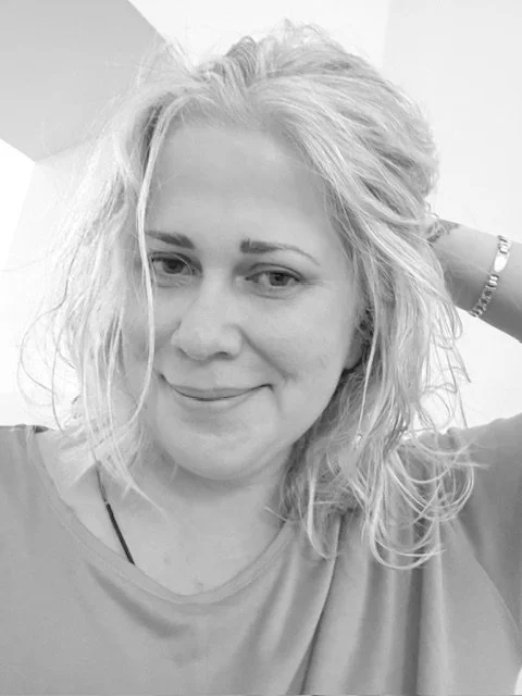 Black and white photo of a woman with wavy blonde hair, smiling softly, wearing a casual t-shirt and a bracelet on her wrist, with her hand resting behind her head.
