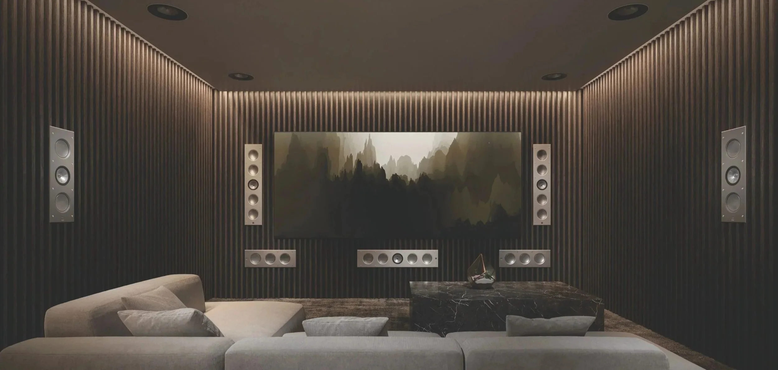 Sala de cine en casa con paredes de madera oscura, un televisor en el centro mostrando una escena de paisaje montañoso, altavoces distribuidos alrededor, sillas y sofá gris en el frente, mesa de centro de mármol negro.