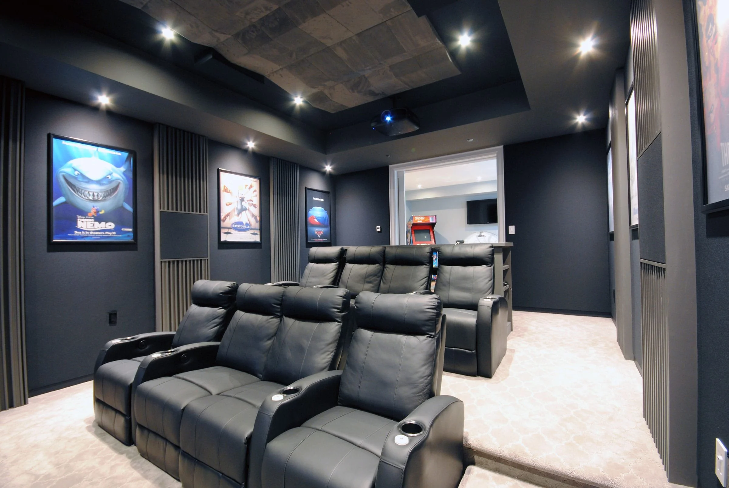 Sala de cine con sillones de cuero negros, paredes grises con posters de películas, y una pantalla en una pared adicional.