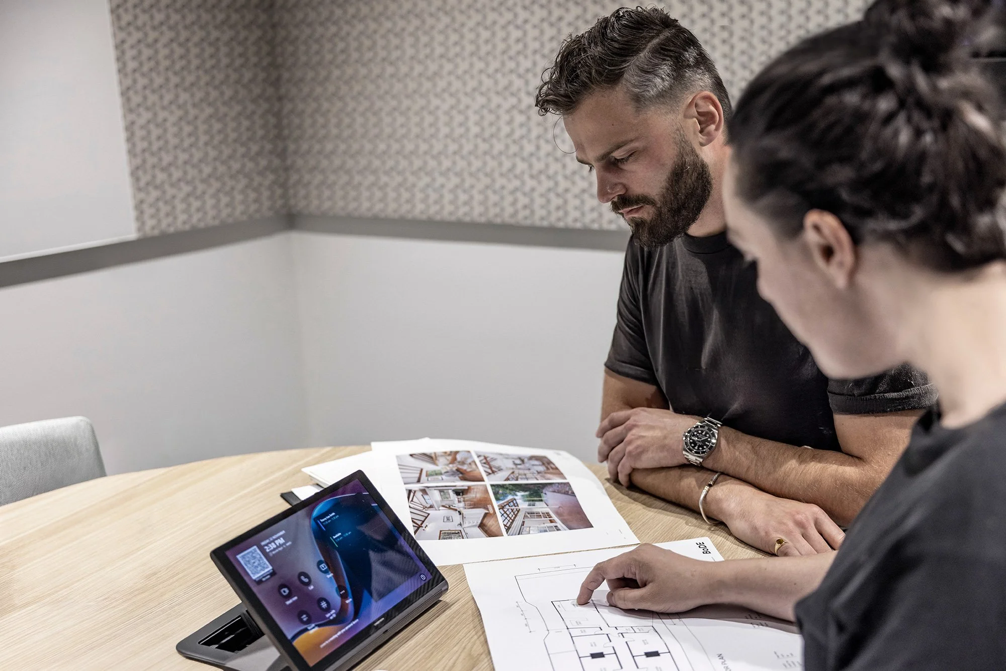 Dos personas revisando planos y materiales de diseño en una mesa de trabajo, con dispositivos electrónicos y revistas de inspiración, trabajando en conjunto para hacer realidad un proyecto.