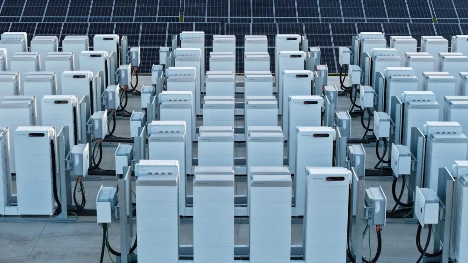 Filas de módulos de baterías eléctricas conectadas en un entorno industrial, con paneles solares en el fondo.