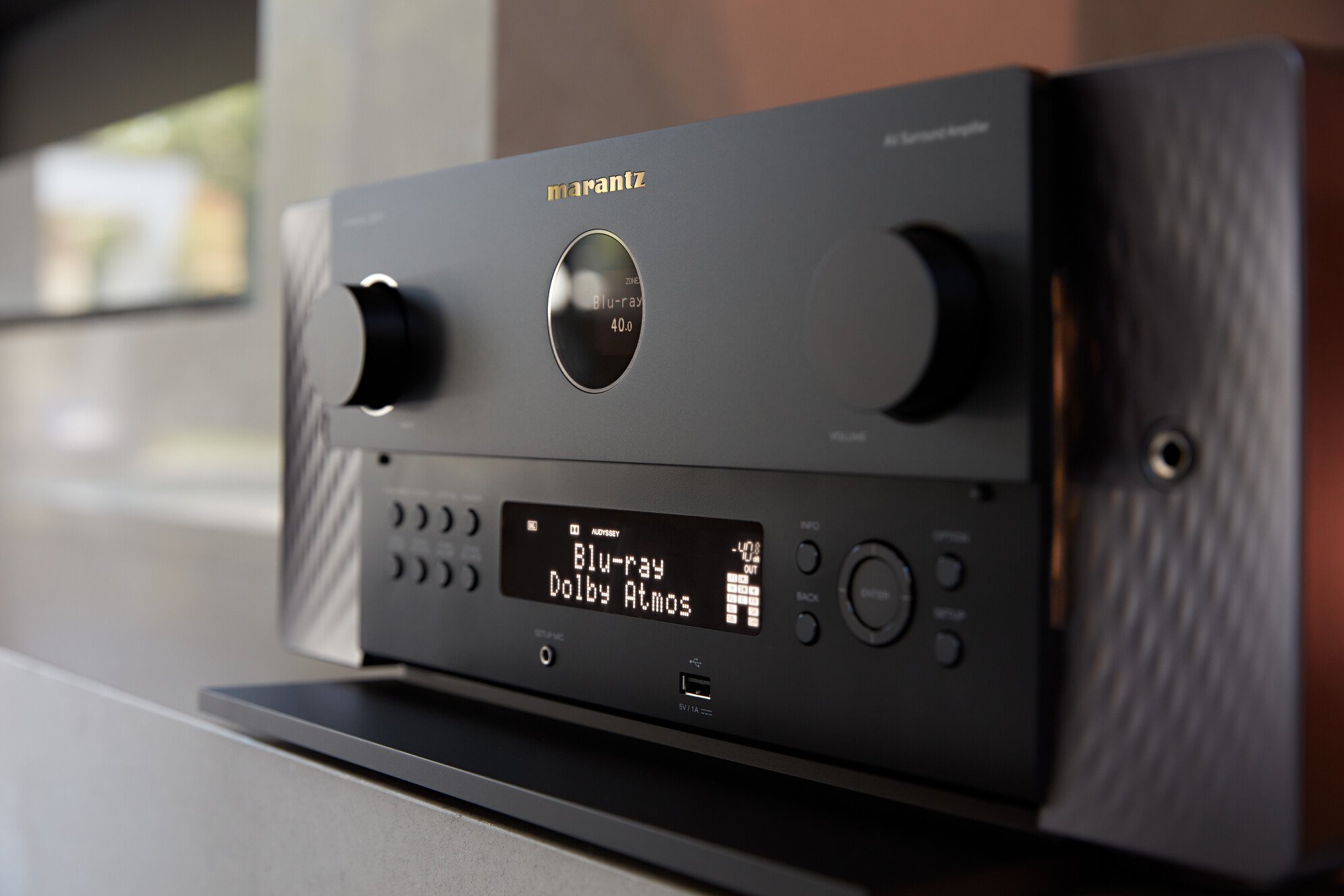 Amplificador de audio negro con pantalla digital que muestra 'Blu-ray Dolby Atmos' y controles en el frente, colocado sobre una mesa con fondo difuso.