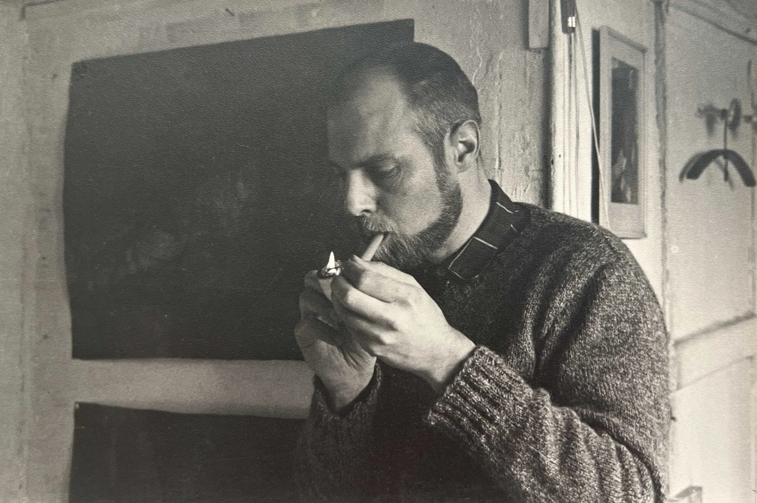 A man with short dark hair and a beard smokes a cigarette while sitting on a bench in a room with exposed brick walls and framed pictures hanging in the background.