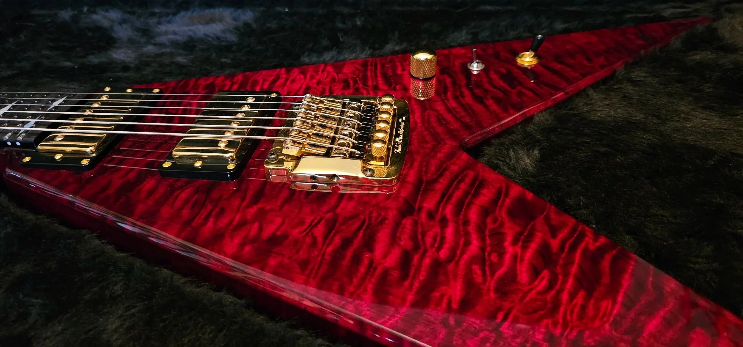 Close-up of a red electric guitar with quilt maple and gold hardware, pickups, and a bridge, lying on a black furry surface.
