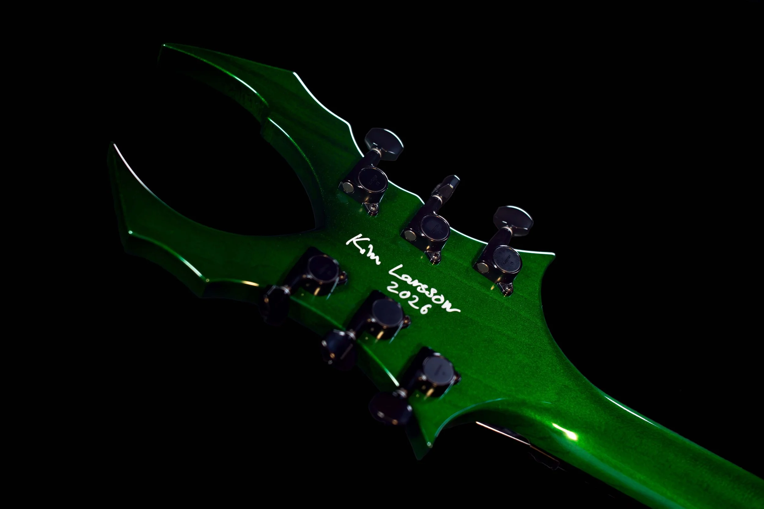 Close-up of a green electric guitar neck with tuning pegs, with handwritten text 'Kim Lawson 2026' on the headstock, against a black background.