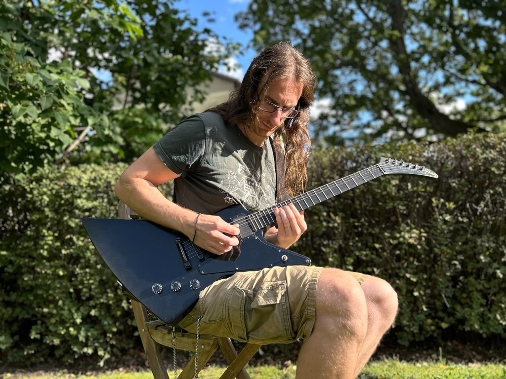 A person sitting outdoors on a wooden chair, playing a black electric guitar with a pointed body shape, against a background of green trees and bushes.
