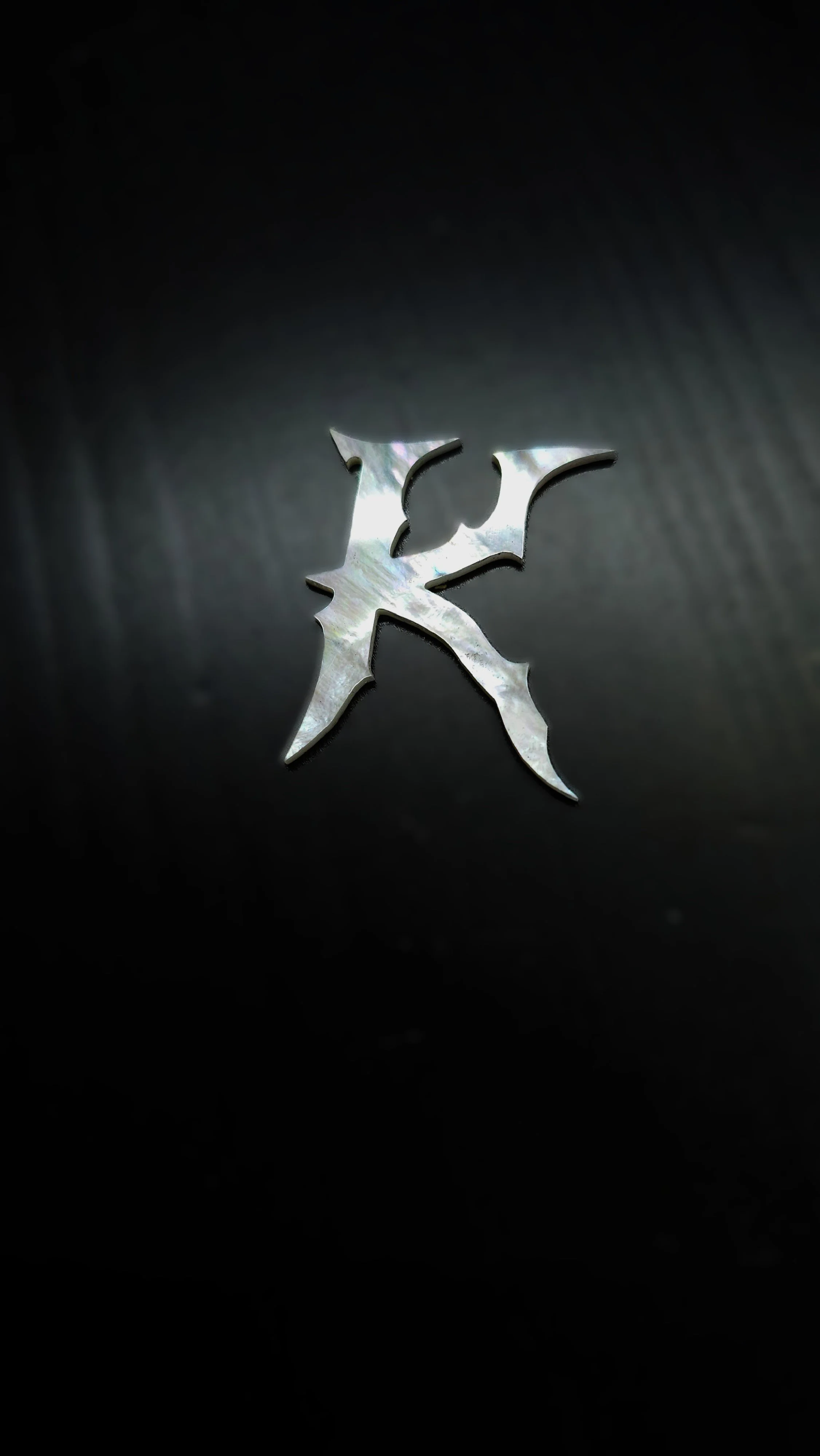 A mother of pearl letter 'K', placed on a dark surface with dramatic lighting that creates reflections and shadows.
