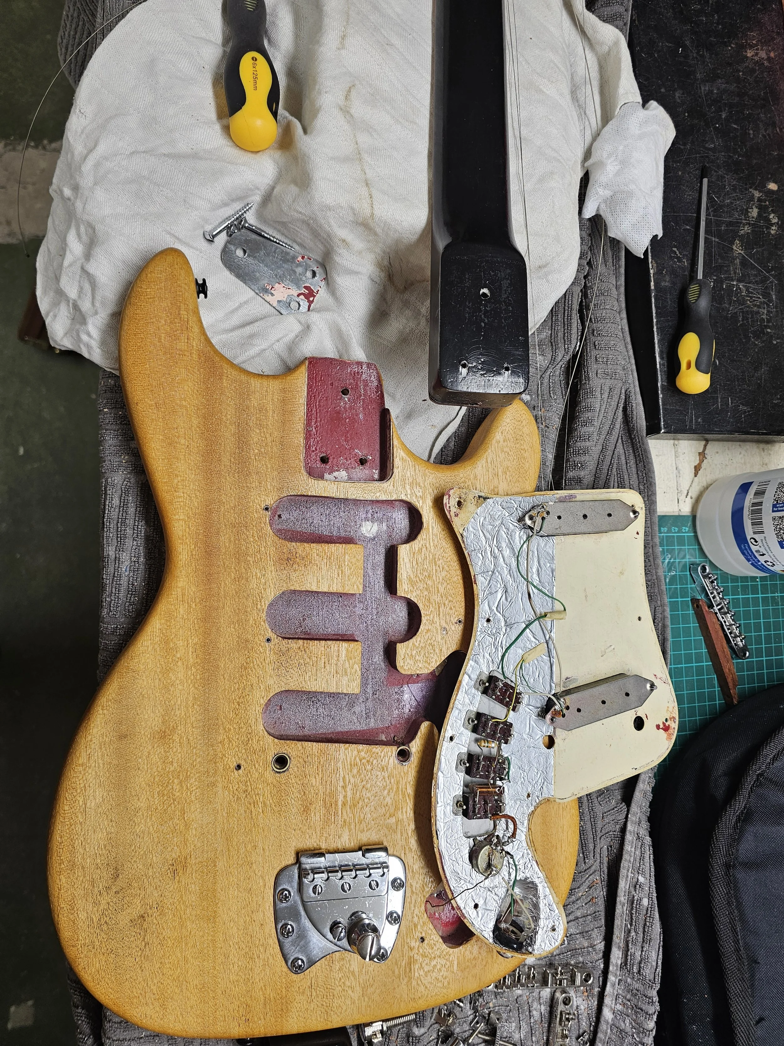 Disassembled electric guitar with its body, neck, and internal wiring visible. Tools and parts such as screwdrivers, screws, and electronic components are scattered around on a work surface.