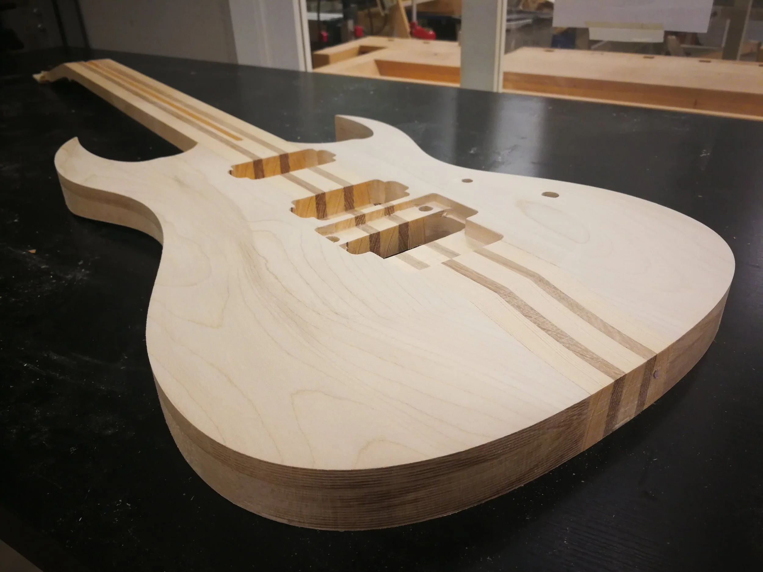 unfinished electric guitar body on a black work table in a woodworking shop, with wooden and woodworking tools in the background.