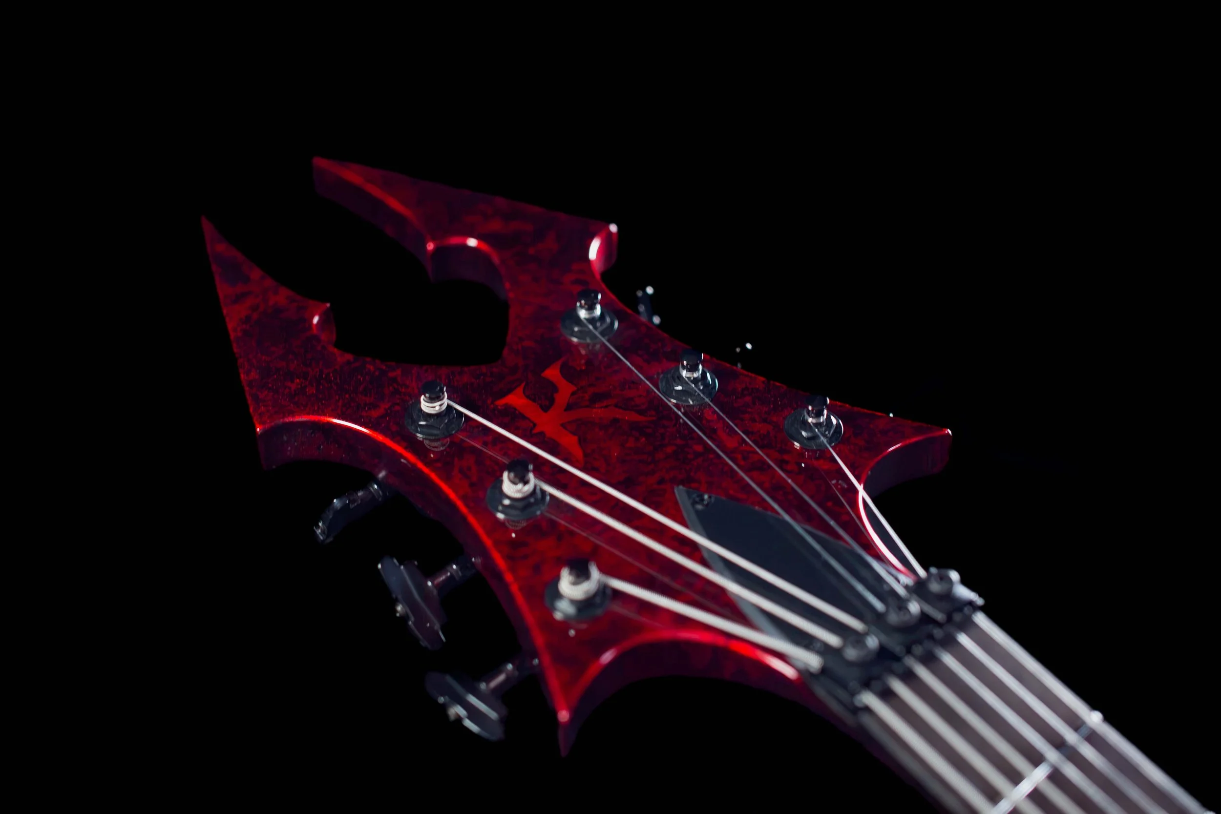 Close-up view of a red electric guitar headstock with unique and aggressive shape, with black hardware, strings, and tuning pegs, against a black background.