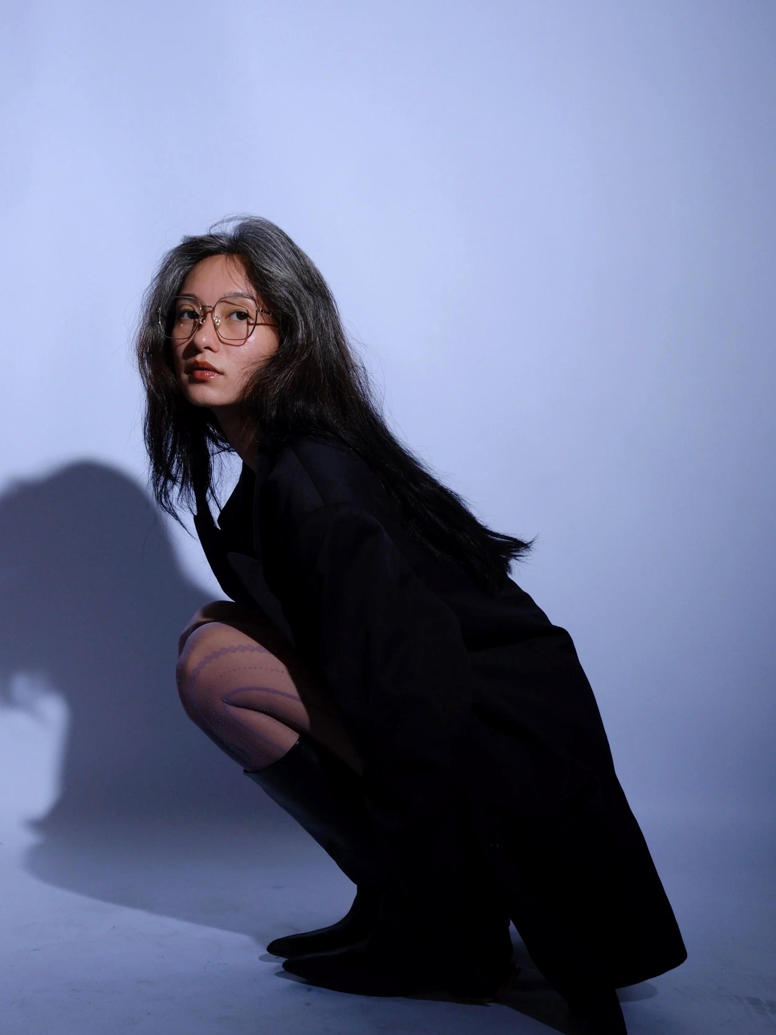 A woman with long dark hair, wearing glasses and black clothing, crouching against a plain light blue background with her head turned slightly to the side.