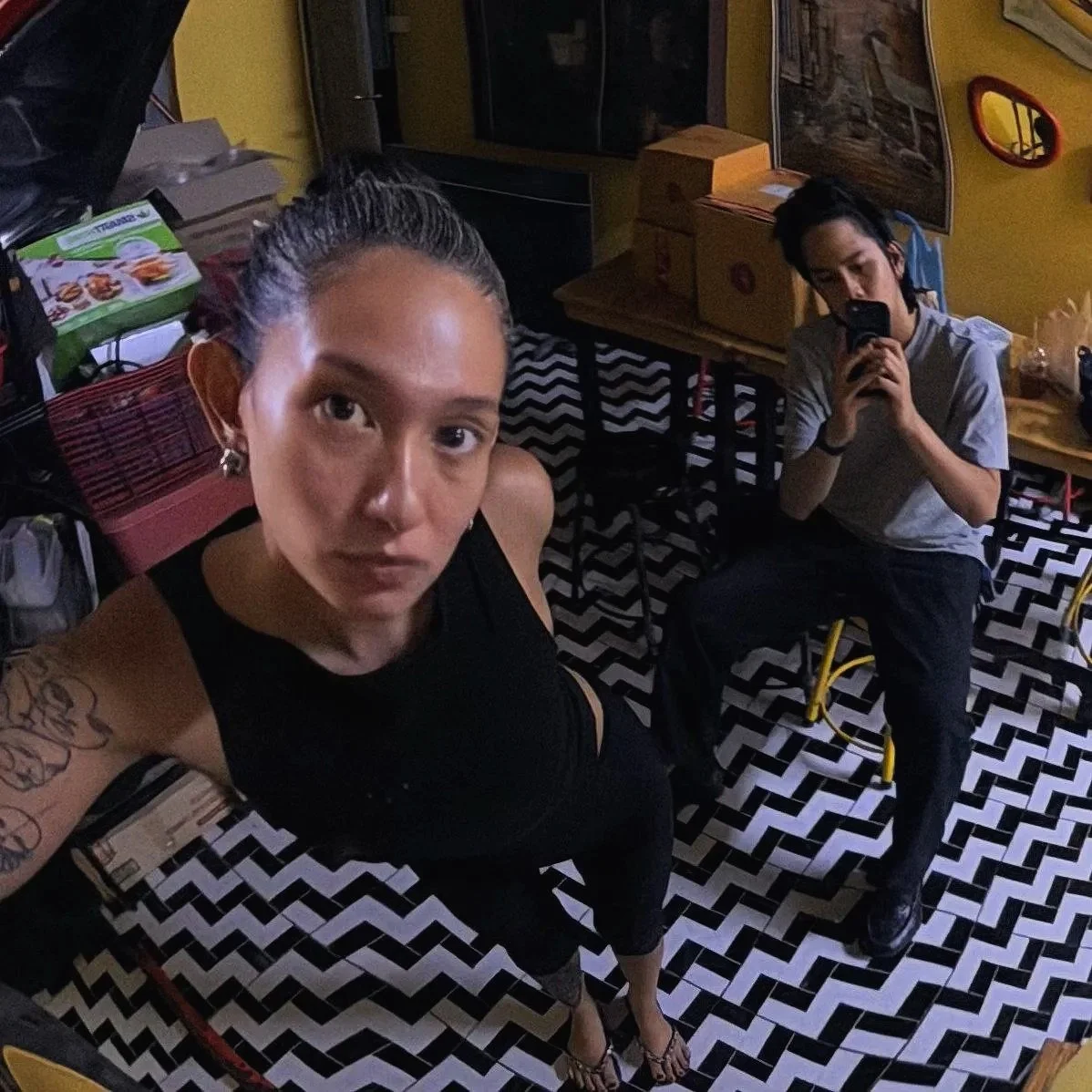 A woman with braided hair, tattoos, and wearing a black top, taking a selfie in a patterned room. A young man with dark hair, wearing a grey shirt, is sitting and looking at his phone in the background.