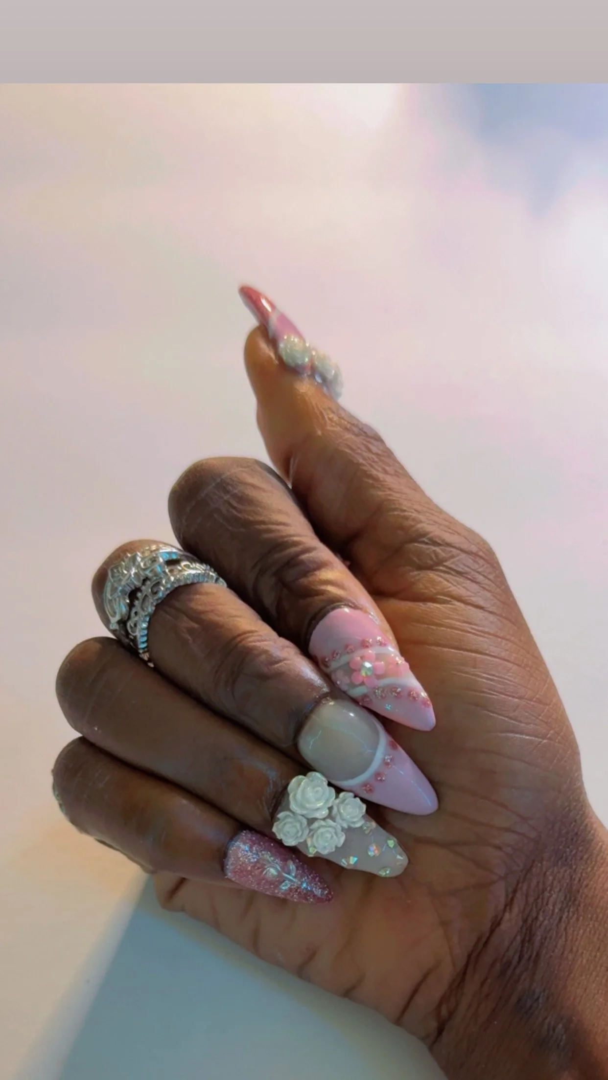 Close-up of a hand with multiple decorated nails and rings, showing pink and white pastel nail designs with floral and glitter accents.