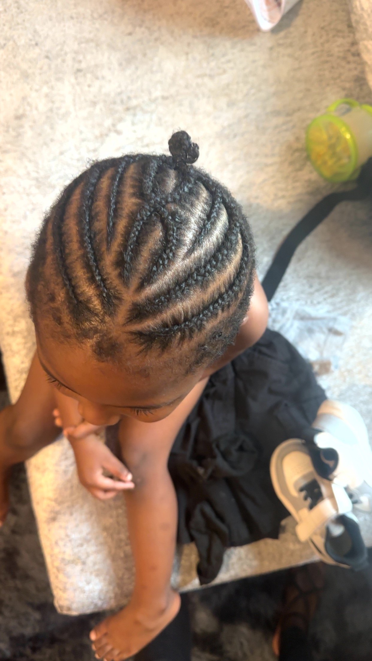 A top view of a child sitting on a bench with their head bowed. The child's hair is styled in cornrows with a small bun on top.