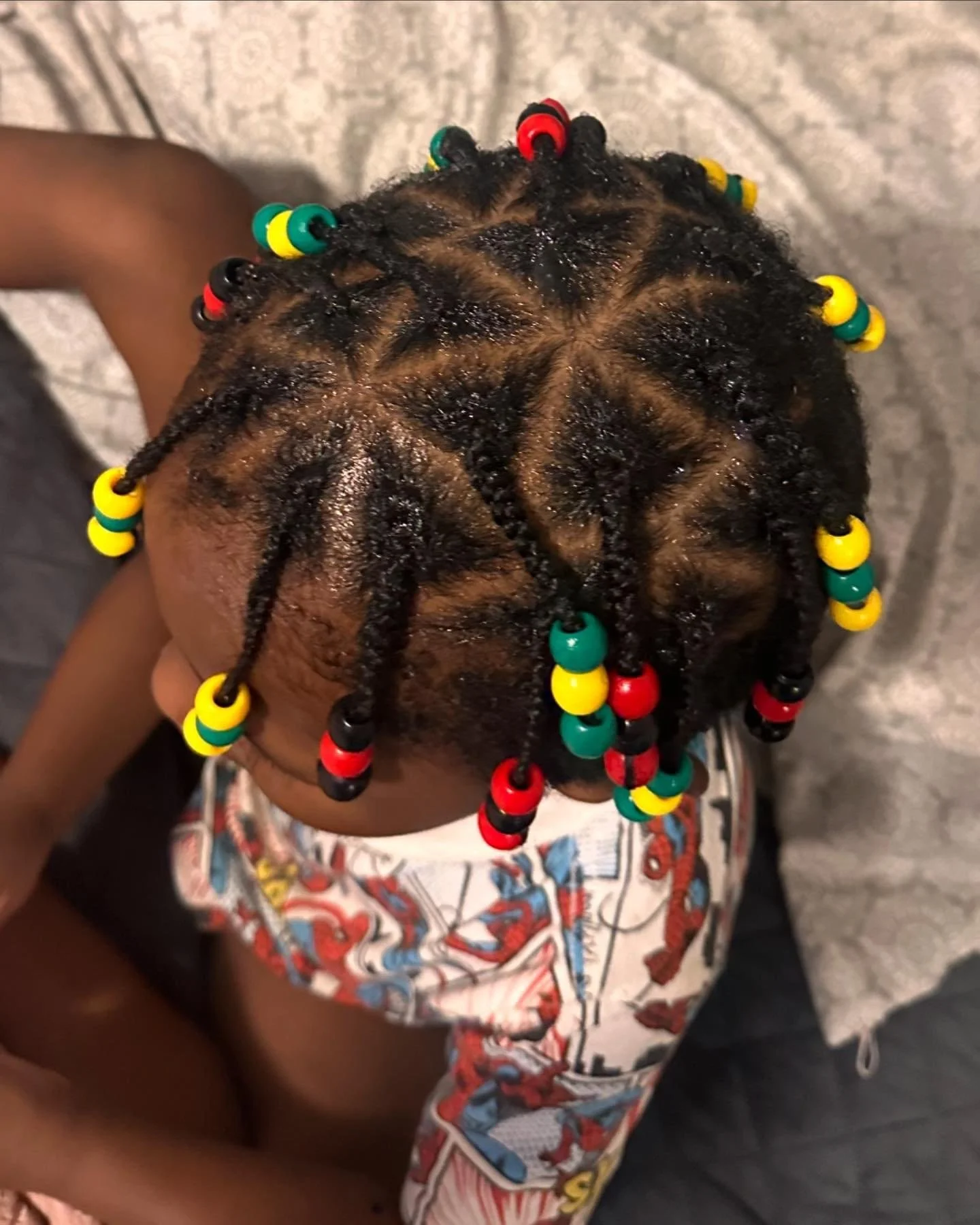 Top view of a young child's head with hair styled in small twists decorated with colorful beads in red, yellow, green, and black. The child is wearing a shirt featuring a superhero pattern.